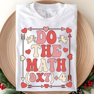 May include: White t-shirt with a retro design that says "DO THE MATH (9X7)+4" in red, pink, and cream colors. The design includes arrows, hearts, and cherubs. The shirt is folded on a wooden tray.