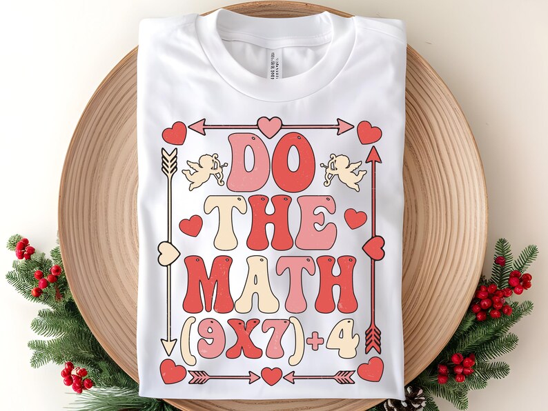 May include: White t-shirt with a retro design that says "DO THE MATH (9X7)+4" in red, pink, and cream colors. The design includes arrows, hearts, and cherubs. The shirt is folded on a wooden tray.