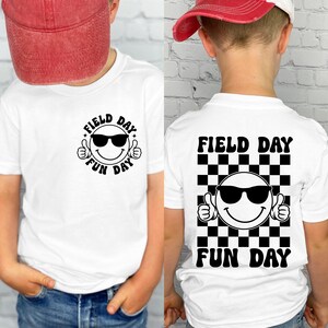 Puede incluir: Camiseta blanca con un gráfico en blanco y negro. La parte delantera presenta una cara sonriente con gafas de sol con las palabras "FIELD DAY" y "FUN DAY". La parte trasera tiene el mismo diseño sobre un fondo a cuadros.