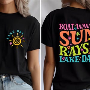 May include: Black t-shirt with a colorful graphic design. The front features a sun with the words "LAKE DAY". The back displays "BOAT WAVES SUN RAYS LAKE DAYS" in vibrant colors.