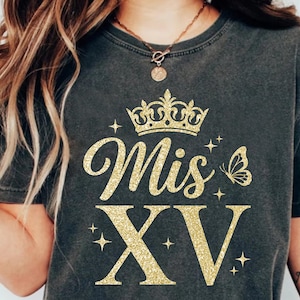 May include: Dark gray t-shirt with gold glitter text and graphics. The text reads "Mis XV" with a crown, butterfly, and stars. A tattoo on the arm reads "More Ocean".