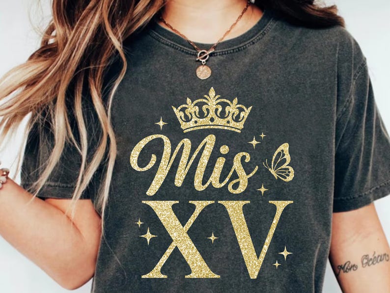 May include: Dark gray t-shirt with gold glitter text and graphics. The text reads "Mis XV" with a crown, butterfly, and stars. A tattoo on the arm reads "More Ocean".