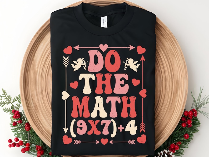 May include: Black t-shirt with a retro design that says "Do The Math (9x7)+4". The design includes red and pink hearts, arrows, and cherubs. The shirt is displayed on a wooden tray with holiday decorations.