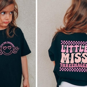 May include: Black t-shirt with a pink graphic on the front featuring a smiley face with heart eyes and peace signs. The back of the shirt has the words "LITTLE MISS THREENAGER" in pink with a checkerboard pattern.