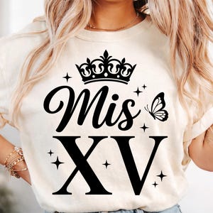 May include: Cream-colored t-shirt featuring the text "Mis XV" in a black script font, with a crown above the word "Mis" and a butterfly to the right. The Roman numeral "XV" is in a bold, blocky font. Small star graphics are scattered around the text.