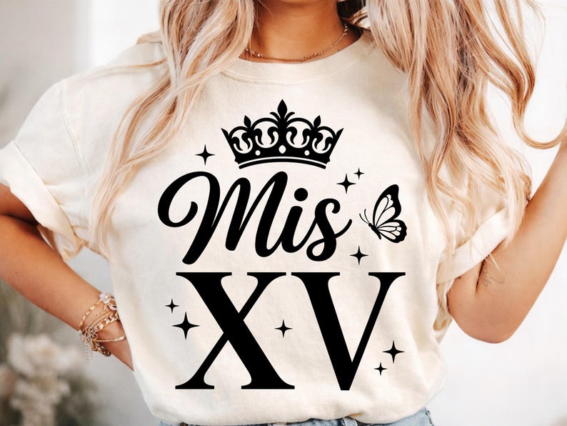 May include: Cream-colored t-shirt featuring the text "Mis XV" in a black script font, with a crown above the word "Mis" and a butterfly to the right. The Roman numeral "XV" is in a bold, blocky font. Small star graphics are scattered around the text.