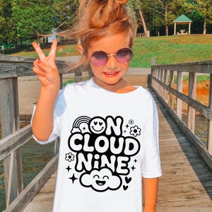 以下が含まれることがあります： 黒い文字で「ON CLOUD NINE」と書かれた白いTシャツ。虹、笑顔、花、星、雲が描かれています。丸いサングラスをかけ、ピースサインをしています。