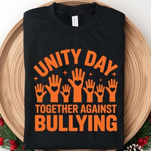 May include: Black t-shirt with orange text and graphics. The design includes the words "UNITY DAY" above a graphic of hands with hearts, and the phrase "TOGETHER AGAINST BULLYING" below. The shirt is displayed on a wooden tray.