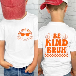 May include: White t-shirts with orange graphics. The front of the shirt features a smiling face wearing sunglasses with the words "BE KIND BRUH". The back of the shirt has the words "BE KIND BRUH" with thumbs up and a checkered pattern.