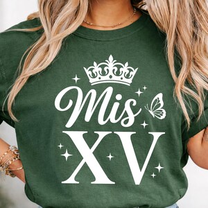 May include: Dark green t-shirt with the words "Mis XV" in white script and block letters, topped with a crown and butterfly graphic. The design includes star accents, creating a festive look.