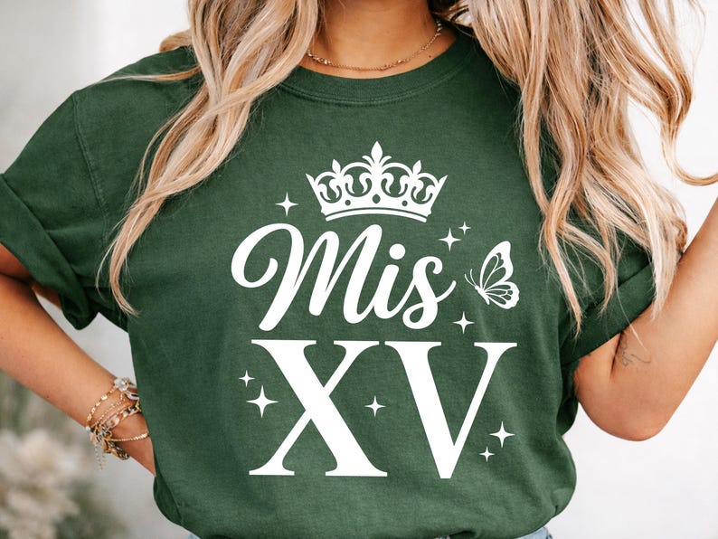May include: Dark green t-shirt with the words "Mis XV" in white script and block letters, topped with a crown and butterfly graphic. The design includes star accents, creating a festive look.