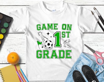 May include: White t-shirt with a green and black graphic that says "GAME ON 1ST GRADE". The design includes a soccer ball, soccer cleats, and a flag. Other items in the image include a watercolor paint set, an orange, and a pair of scissors.