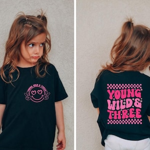 May include: Black t-shirt with the text "Young Wild & Three" in pink. The front features a smiley face with peace signs and hearts. The back has the same text with a checkered pattern.