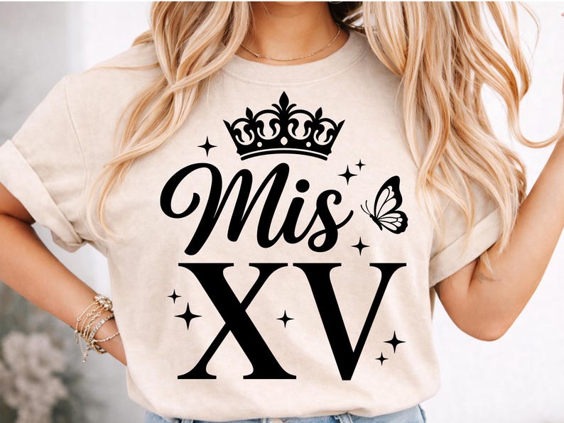 May include: Beige t-shirt with a black crown, the words "Mis XV", a butterfly, and the Roman numeral XV. The design includes star accents. The shirt has short sleeves.
