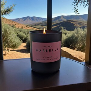 Op de afbeelding: Een zwarte kaars met een roze label waarop staat "We are Marbella Concha Lemongrass & Ginger". De kaars is aangestoken en de vlam is zichtbaar. De kaars staat op een houten oppervlak met uitzicht op bergen en bomen op de achtergrond.