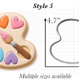 Op de afbeelding: Een paletvormig koekje versierd met zes kleurrijke hartvormige glazuurvlekken en een penseel, naast een metalen koekjesvorm op een rasterachtergrond, tekst Style 5, 11,9 cm bij 12,7 cm, en Multiple sizes available.