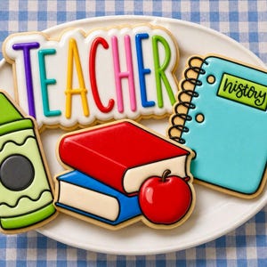 Op de afbeelding: Een verzameling versierde koekjes op een wit bord, waaronder een koekje in de vorm van een waskrijtje, een stapel van twee boeken met een appel, een spiraalblok met het label history en een koekje met het woord TEACHER in gekleurd glazuur.