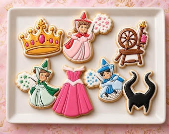 Peut inclure: Un ensemble de sept biscuits décorés sur un plateau rectangulaire blanc. Les formes incluent une couronne dorée, une fée en rose avec une baguette, un rouet marron, une fée en vert, une robe rose, une fée en bleu et des cornes noires.