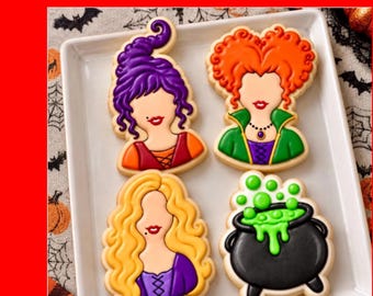 Op de afbeelding: Vier versierde koekjes op een wit vierkant bord. Drie koekjes hebben de vorm van vrouwelijke figuren met opvallende kapsels in paars, oranje en geel, en één koekje heeft de vorm van een zwarte ketel met felgroene borrelende vloeistof.