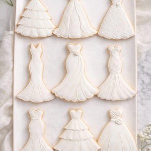 May include: A collection of nine white dress-shaped sugar cookies arranged on a rectangular tray. Each cookie features intricate royal icing details, including tiered ruffles, lace textures, floral accents, and various silhouettes like mermaid and A-line.