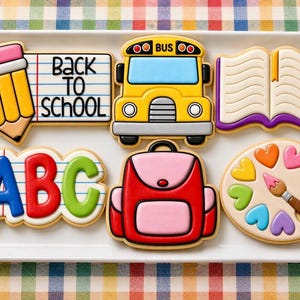 Op de afbeelding: Een set van zes schoolthema koekjes op een witte rechthoekige schaal. De koekjes hebben de vorm van een potlood met de tekst Back To School, een gele schoolbus, een open boek, de letters ABC, een rode en roze rugzak en een verfpalet.