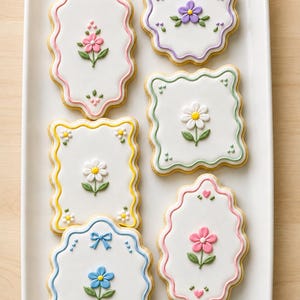 May include: Six rectangular and oval sugar cookies with scalloped edges arranged on a white rectangular platter. Each cookie features a smooth white icing base decorated with colorful piped royal icing flowers, small green leaves, and dots.
