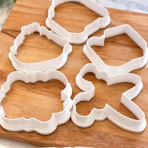 Op de afbeelding: Een set van vier witte plastic koekjesvormen in verschillende vormen, waaronder een afgerond vierkant, een geschulpte rechthoek, een eenvoudige rechthoek en een kruisvorm. De tekst "Sample set of cookie cutters" staat onderaan de afbeelding.