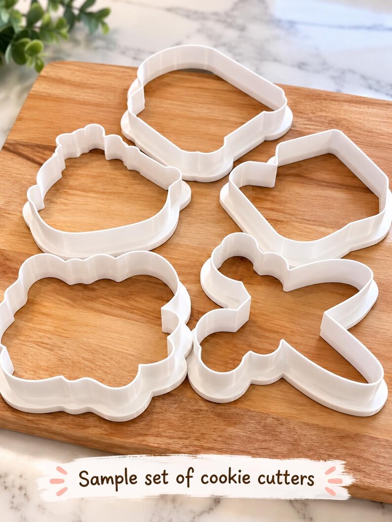 Op de afbeelding: Een set van vier witte plastic koekjesvormen in verschillende vormen, waaronder een afgerond vierkant, een geschulpte rechthoek, een eenvoudige rechthoek en een kruisvorm. De tekst "Sample set of cookie cutters" staat onderaan de afbeelding.