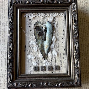 May include: Framed mixed-media artwork featuring a distressed metal heart with a key, set against a backdrop of sheet music and lace. The word "LOVE" is spelled out at the bottom with individual letter tiles. The frame is dark with ornate detailing.