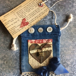 May include: A denim pouch with a heart-shaped embellishment and a small padlock. The pouch has three white buttons and a lace trim. A vintage tag with text from "Antony and Cleopatra" is attached.