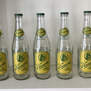 May include: Five clear glass Vernor's ginger soda bottles with green caps. Each bottle has a yellow label with the Vernor's logo and text. The bottles are 12 fl oz (355 ml) and are a 2000 Limited Edition.