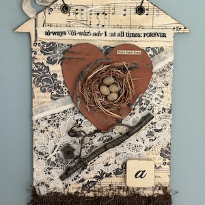 May include: A mixed-media wall hanging in the shape of a house. It features a heart with a nest and eggs, lace, sheet music, and the text "always at all times: FOREVER". A key and a small wooden block with the letter "a" are also included.