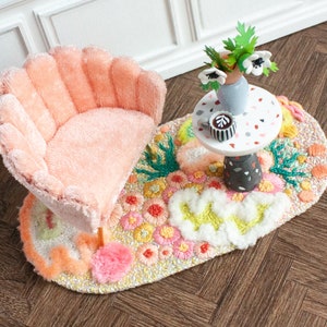 May include: A pink, plush, scalloped armchair sits on a colorful, oval rug with a small, white table with a cup of coffee and a vase of white flowers.