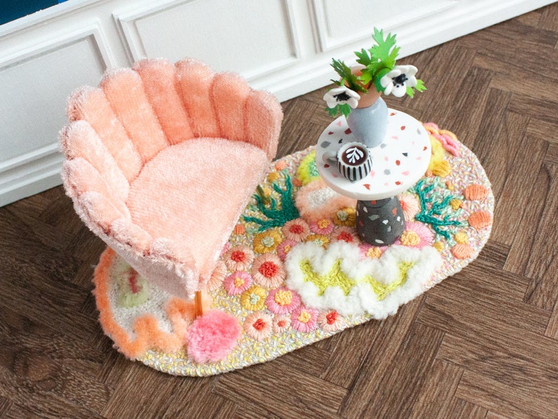 May include: A pink, plush, scalloped armchair sits on a colorful, oval rug with a small, white table with a cup of coffee and a vase of white flowers.