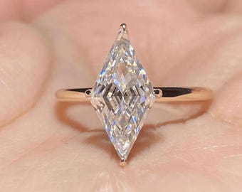 Pode incluir: Um anel de noivado em forma de diamante com uma gema grande, clara e facetada. O anel tem uma banda fina de ouro. A gema é definida com uma parte superior e inferior pontiagudas. O anel é mostrado contra um fundo rosa suave.