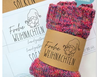 Op de afbeelding: Een kleurrijke, handgebreide sok met een bruine papieren band met de tekst "Frohe Weihnachten" en een kabouterillustratie. De sok is een mix van roze, paars en gele wol. Extra papier met tekst is zichtbaar op de achtergrond.