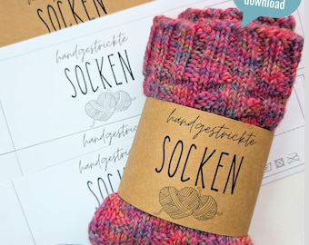 Op de afbeelding: Handgebreide sokken in roze, paarse en blauwe tinten, omwikkeld met een bruine papieren band met de tekst "handgestrickte SOCKEN". Extra labels met dezelfde tekst en een spraakbel "digitaler download" zijn ook zichtbaar.