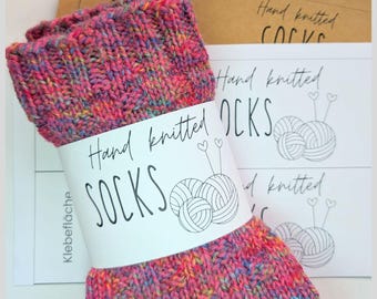 Op de afbeelding: Handgebreide sokken in levendige roze, gele en blauwe tinten. De sokken zijn verpakt met een label met de tekst "Hand knitted SOCKS" met een afbeelding van bollen wol en breinaalden. Een digitale download label is ook zichtbaar.