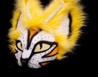 May include: A yellow fur cat mask with black and orange stripes, white spots, and yellow eyes. The mask has a white nose and mouth.