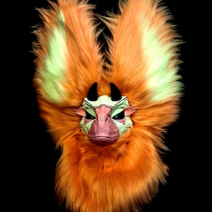 May include: A creature mask featuring long, vibrant orange faux fur with light green highlights. The central face is textured, light green with a pinkish snout, two small black horns, and narrow, dark, almond-shaped eye openings.