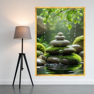 Op de afbeelding: Een ingelijste print toont een zen-tuin scène. Gladde, grijze stenen zijn gestapeld, met groen mos en waterdruppels. De achtergrond toont weelderig groen en een wateroppervlak, omlijst in goud.