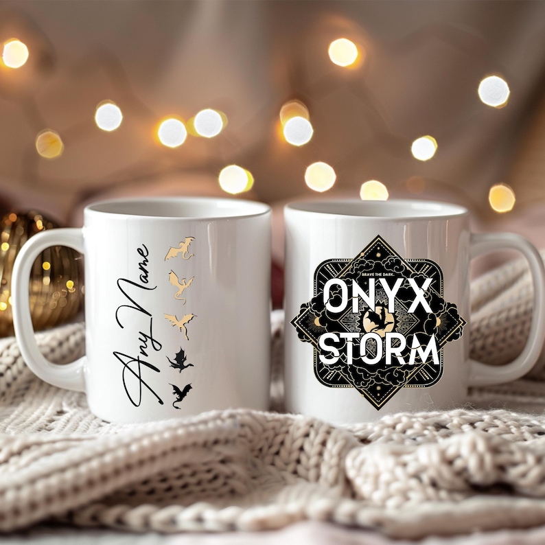 K&ouml;nnte beinhalten: Zwei wei&szlig;e Keramikbecher. Ein Becher zeigt ein schwarzes und goldenes Design mit dem Text "ONYX STORM" und der andere Becher hat ein goldenes Design mit f&uuml;nf Drachen und dem Text "Any Name".