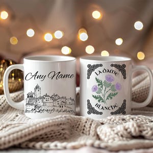 May include: Two white ceramic mugs. One mug features the text "Any Name" above a black and white illustration of a village. The other mug has the text "La Dame Blanche" with a purple thistle design and Celtic knot accents.