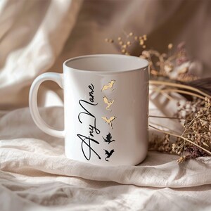 K&ouml;nnte beinhalten: Wei&szlig;e Keramiktasse mit einem schwarzen und goldenen Drachenmotiv und dem Text "Any Name" in schwarzer Kursivschrift.