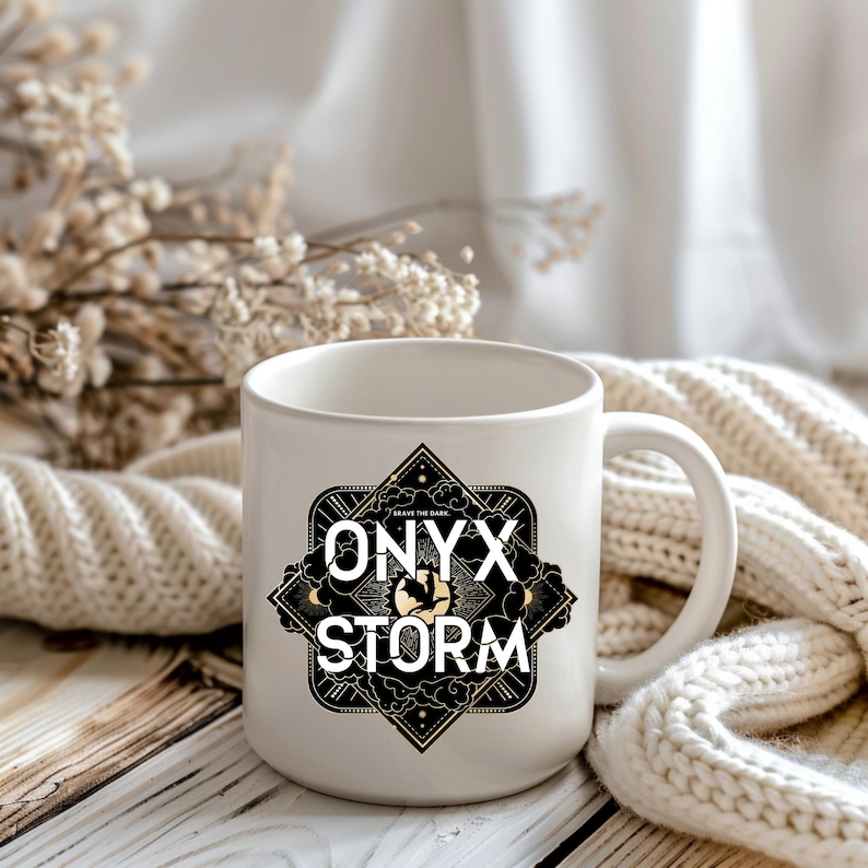K&ouml;nnte beinhalten: Wei&szlig;e Keramiktasse mit einem schwarzen und goldenen Design. Das Design zeigt ein geometrisches Muster mit den Worten "Onyx Storm" und dem Text "Brave the Dark".