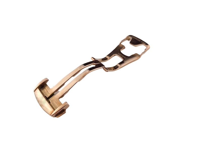 May include: A rose gold watch clasp, featuring a curved design with a hinged mechanism. The clasp is made of a shiny, metallic material and is designed to secure a watch band. The overall shape is elongated and slightly curved.