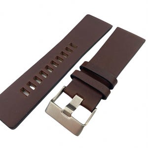 Puede incluir: Una correa de reloj de cuero marrón con una hebilla plateada. La correa tiene una serie de agujeros para ajustar el tamaño.