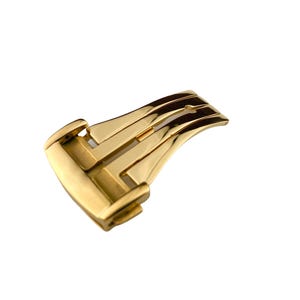 May include: A gold-colored watch clasp, featuring a curved, rectangular design with multiple parallel grooves. The clasp has a polished finish, reflecting light, and is set against a white background.