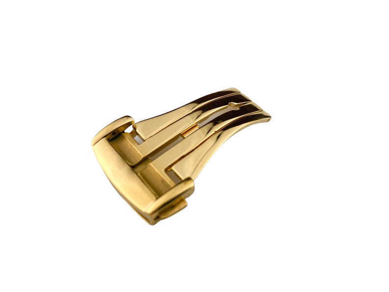 May include: A gold-colored watch clasp, featuring a curved, rectangular design with multiple parallel grooves. The clasp has a polished finish, reflecting light, and is set against a white background.