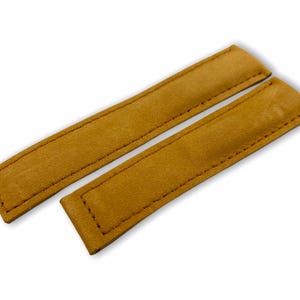 Puede incluir: Dos correas de reloj de cuero color mostaza. Cada correa es rectangular con extremos redondeados y bordes cosidos. Las correas están hechas de cuero suave y texturizado sobre un fondo blanco.